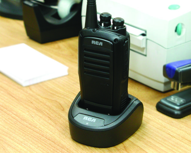 An RCA 2-way handheld radio rests in its charger on a desk.